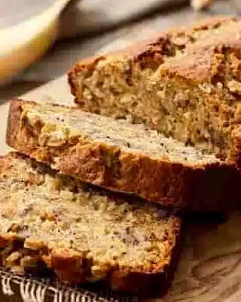 Slices of moist banana bread on a wooden board with ripe bananas in the background